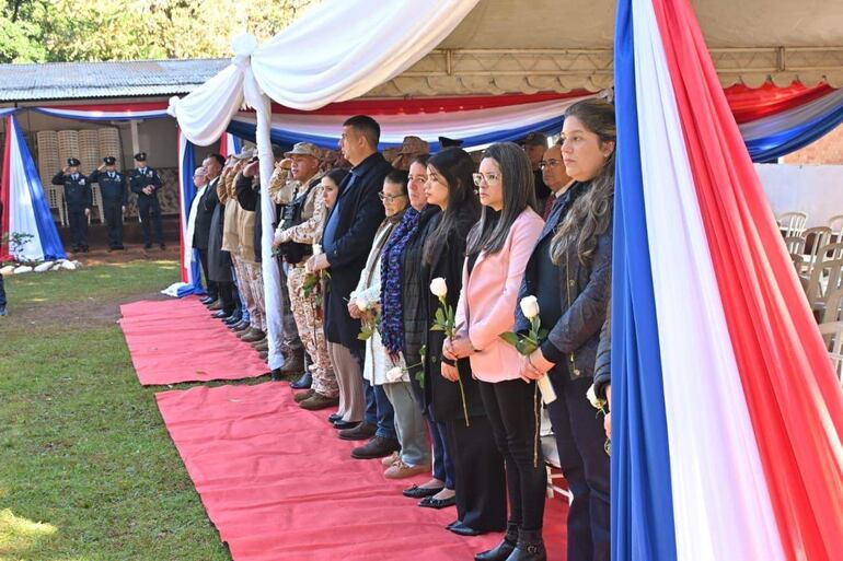 Un grupo de personas en atuendos formales sostiene rosas blancas en un evento con bandera paraguaya de fondo.