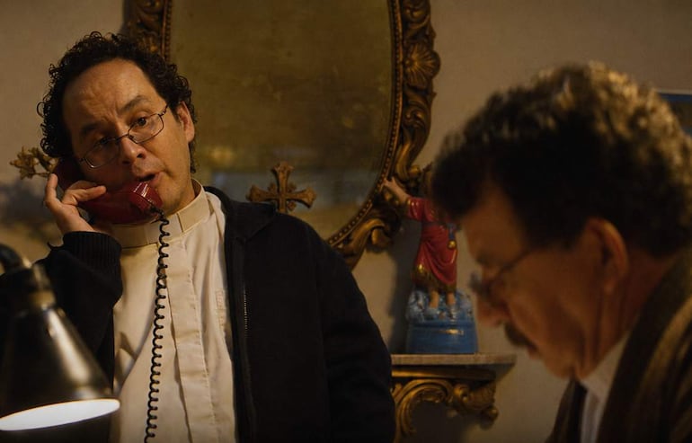 Hombre de cabello rizado y gafas habla por teléfono rojo, mientras otro hombre serio escucha en un despacho con decoraciones religiosas.