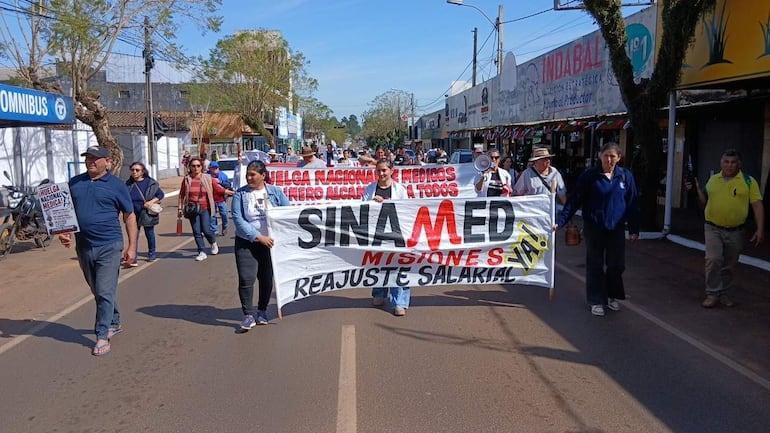 Mujer con megáfono lidera la marcha de médicos en San Ignacio, sosteniendo pancartas sobre reajuste salarial y huelga nacional.