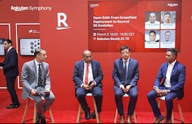 Cuatro hombres en trajes oscuros discuten en evento tecnológico con cartel de Rakuten y fondo rojo.