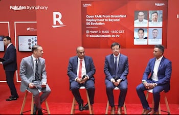 Cuatro hombres en trajes oscuros discuten en evento tecnológico con cartel de Rakuten y fondo rojo.