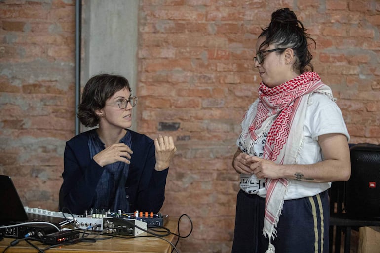Mujer con gafas y abrigo azul habla con énfasis, mientras otra mujer con chal rojo escucha atentamente en ambiente interior.