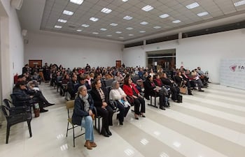 Audiencia de 150 personas en salón iluminado, algunos tomando notas y otros con teléfonos. Ambiente formal y organizado.