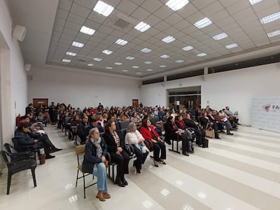 Audiencia de 150 personas en salón iluminado, algunos tomando notas y otros con teléfonos. Ambiente formal y organizado.