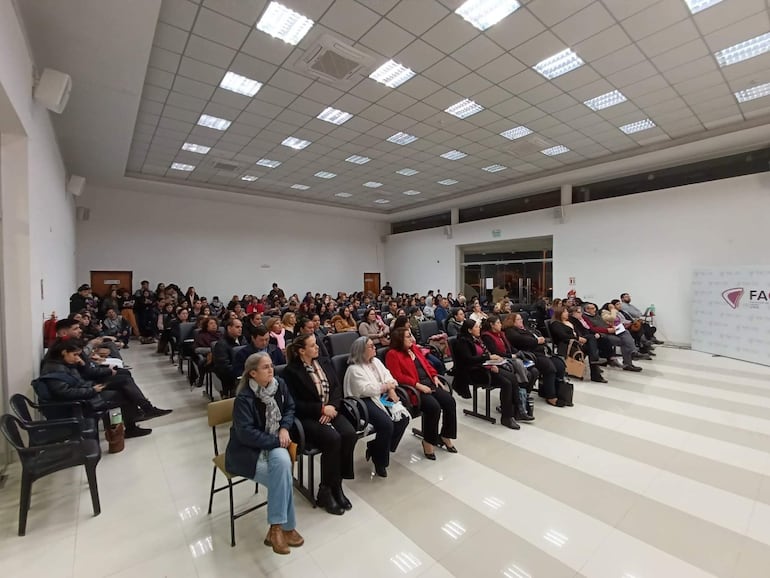 Audiencia de 150 personas en salón iluminado, algunos tomando notas y otros con teléfonos. Ambiente formal y organizado.
