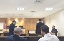 Un hombre de pie discute en la sala del tribunal mientras otros dos hombres observan en actitud neutral.
