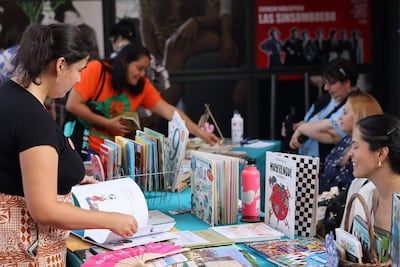 Mujeres en la feria de libros, una observa un libro mientras otra sostiene varios textos en un ambiente acogedor.