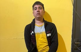 Hombre joven, de pie con manos esposadas, viste camiseta amarilla y gris, abrigo negro y pantalones oscuros en ambiente sencillo.
