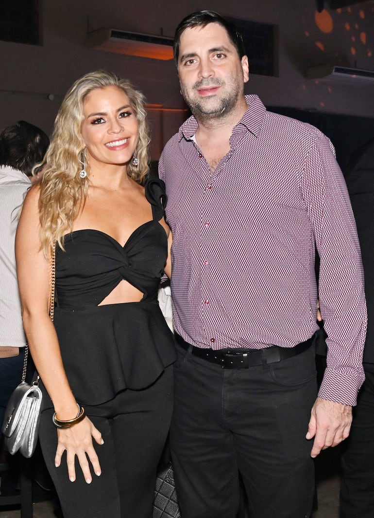 Dustin y una mujer de cabello rubio y rizado sonríen en un evento, ella viste prenda negra y sostiene un bolso plateado.