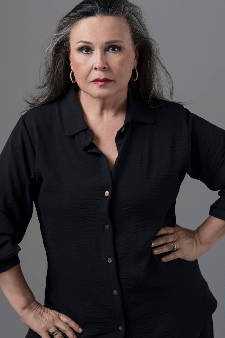 Lizza Bogado, con cabello largo y canoso, vestida con camisa negra, mira seriamente con las manos en las caderas sobre fondo gris.