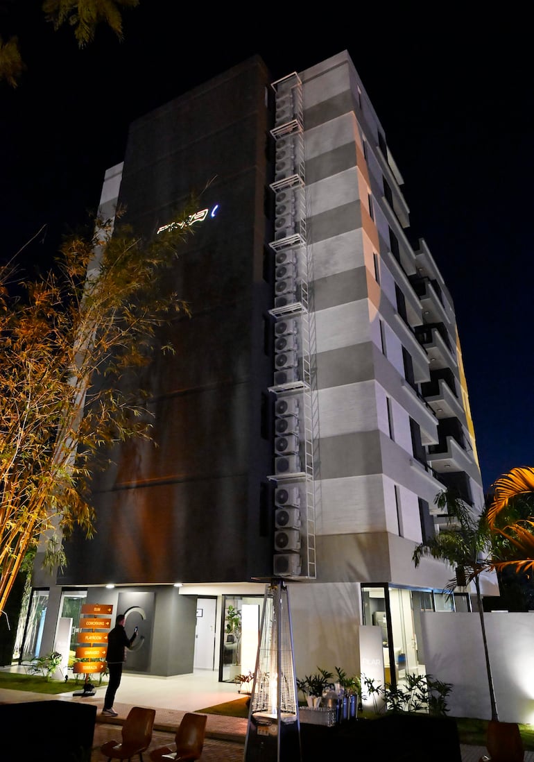 Edificio moderno con fachada gris y detalles en blanco, rodeado de vegetación, iluminado en un ambiente urbano nocturno.