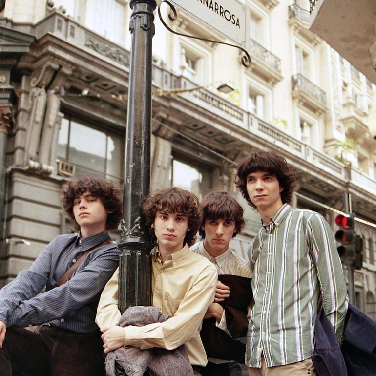 Cuatro jóvenes de cabello rizado conversan, apoyados en un poste de luz, con una chaqueta abrazada y camisetas de diferentes colores.