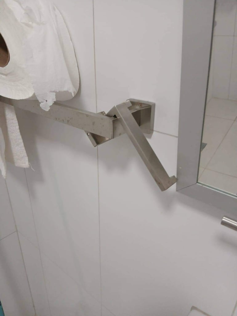 Baño con soporte metálico dañado y toalla de papel blanca. Ambiente interior con paredes de cerámica claras.