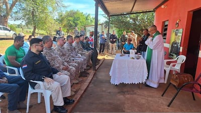 Grupo de personas sentadas, un sacerdote de sotana blanca frente a mesa decorada, entorno rural con árboles.