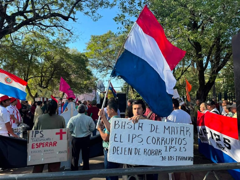 Multitud de manifestantes con banderas paraguayas y pancartas denunciando corrupción del IPS, expresando descontento y urgencia.