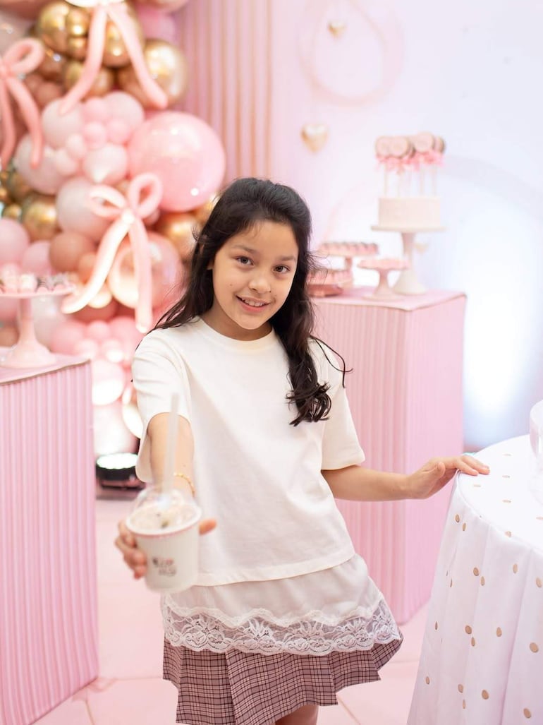 Niña con cabello oscuro, camiseta blanca y falda a cuadros sonríe sosteniendo un vaso en medio de globos rosa y dorado.