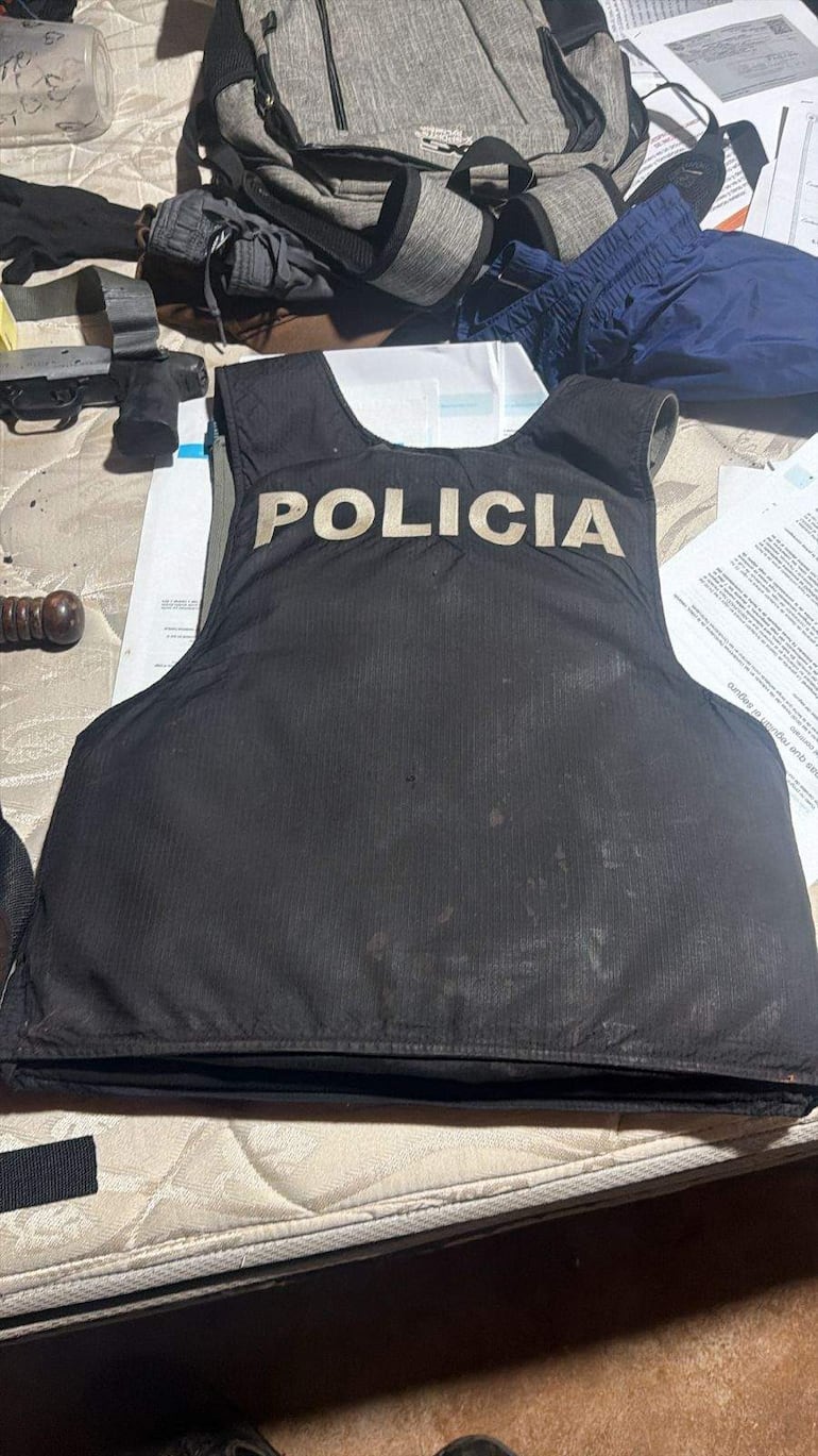 Chaleco antibalas con la palabra 'POLICIA' sobre un colchón, rodeado de ropa, una mochila y un arma de fuego.