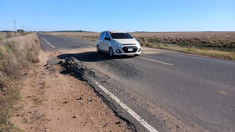 Automóvil blanco circula sobre pavimento agrietado, mientras se observa un paisaje de campos abiertos bajo un cielo despejado.