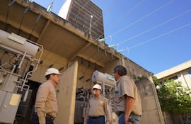 Tres hombres en cascos discuten sobre equipo eléctrico, con transformadores y cableado visibles en un día soleado.