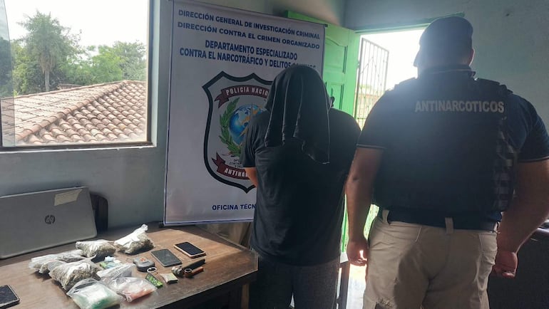 Detenido encapuchado con camiseta negra y pantalones oscuros, mientras un oficial antinarcóticos en camiseta azul supervisa en oficina.