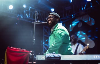 PJ Morton, con chaqueta verde brillante, sonríe mientras toca el piano, acompañado por otro músico en un escenario iluminado.