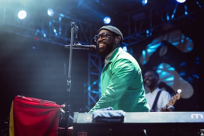 PJ Morton, con chaqueta verde brillante, sonríe mientras toca el piano, acompañado por otro músico en un escenario iluminado.