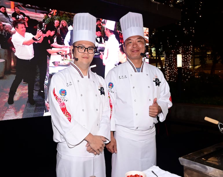 Dos chefs, uno con gafas y serio, y otro amigable con pulgar arriba, posan en evento culinario al aire libre con gran pantalla de fondo.