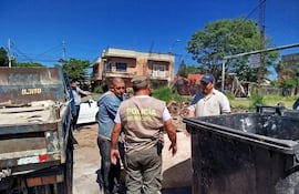 Hombre de camiseta azul observa a un policía municipal hablando, mientras otro hombre con gorra escucha atentamente en un entorno urbano.