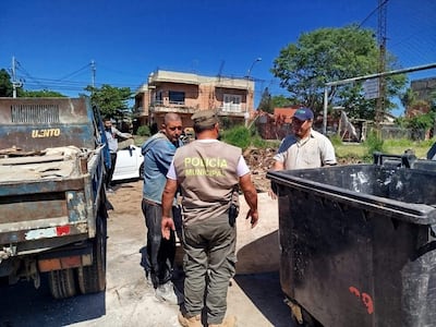 Hombre de camiseta azul observa a un policía municipal hablando, mientras otro hombre con gorra escucha atentamente en un entorno urbano.