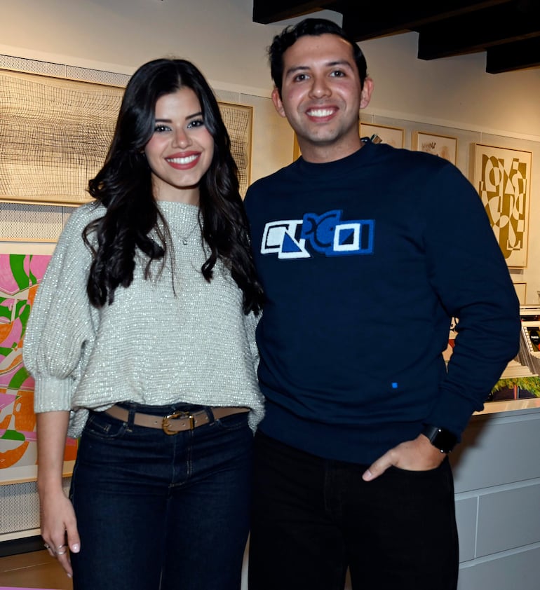 Mujer en suéter claro y hombre en suéter azul sonríen en un ambiente decorativo y cultural, posando juntos.
