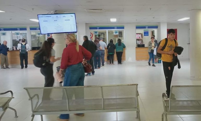 Mujeres conversan en fila, una sostiene un peluche. Lugar luminoso con paredes blancas y suelos de cerámica.