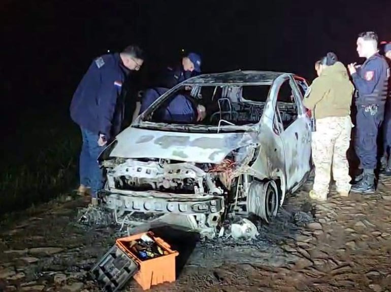 Grupo de cinco personas, algunas en ropa oscura, observan un coche blanco quemado en un área oscura, posiblemente rural.