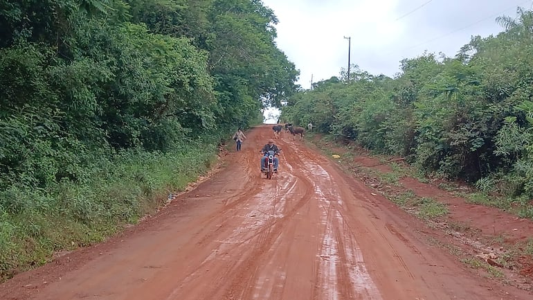 Tres hombres caminan por un camino de tierra; uno en motocicleta y dos a pie, con ambiente nublado y terreno fangoso.