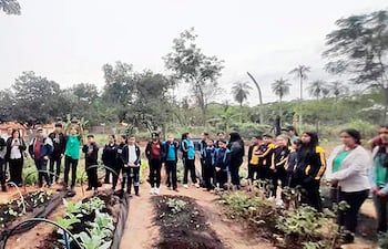 Grupo de 30 personas, incluidos niños y adultos, escuchando atentamente a un orador en un huerto comunitario al aire libre.