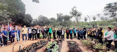 Grupo de 30 personas, incluidos niños y adultos, escuchando atentamente a un orador en un huerto comunitario al aire libre.