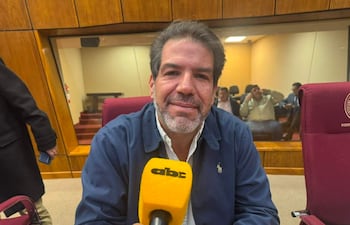 Rodrigo Blanco con chaqueta azul y camisa blanca, sonriendo frente a un micrófono amarillo, en un entorno formal.