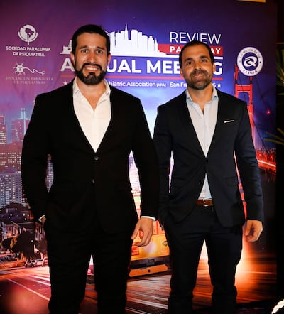 Hombres en trajes formales posan en evento, uno con saco negro y otro con saco azul, en un ambiente decorado con paisaje urbano nocturno.