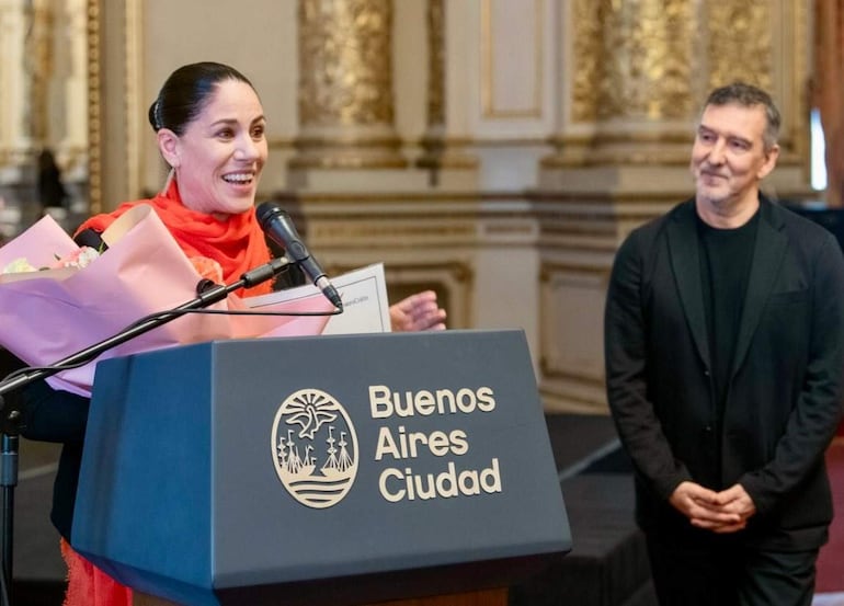 Mujer en blusa negra y abrigo rojo sostiene flores y un documento frente a un micrófono, mientras un hombre la observa con interés.