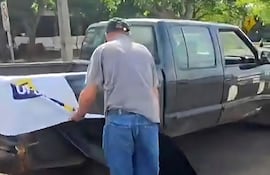 Hombre de espaldas en camiseta gris y jeans, manipula un letrero en camioneta negra, con árboles y señales viales de fondo.