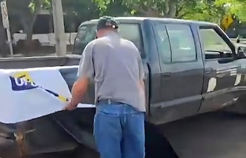 Hombre de espaldas en camiseta gris y jeans, manipula un letrero en camioneta negra, con árboles y señales viales de fondo.