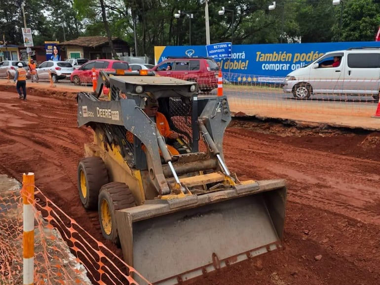 Minicargador John Deere en construcción con un trabajador al volante. Otros obreros en chaquetas naranjas realizan excavación.