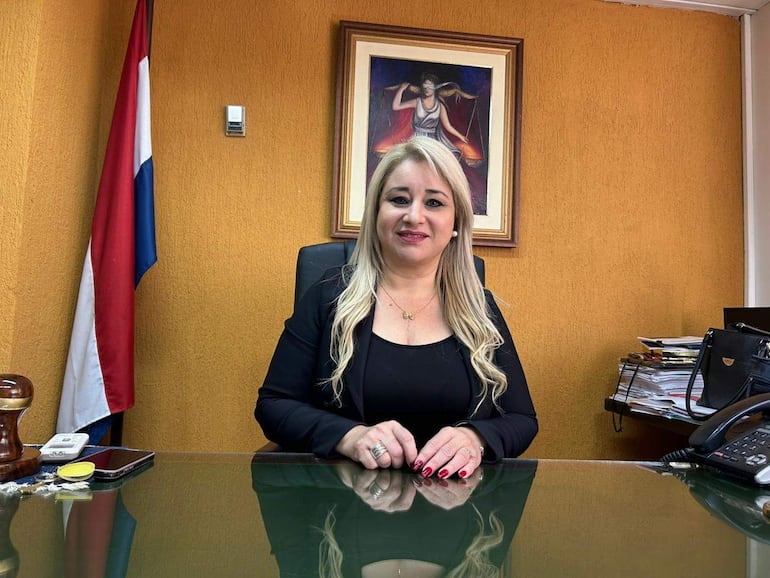 Rosarito Montanía, mujer de cabello rubio y blusa negra, sonríe en su oficina con una bandera paraguaya y documentos sobre el escritorio.