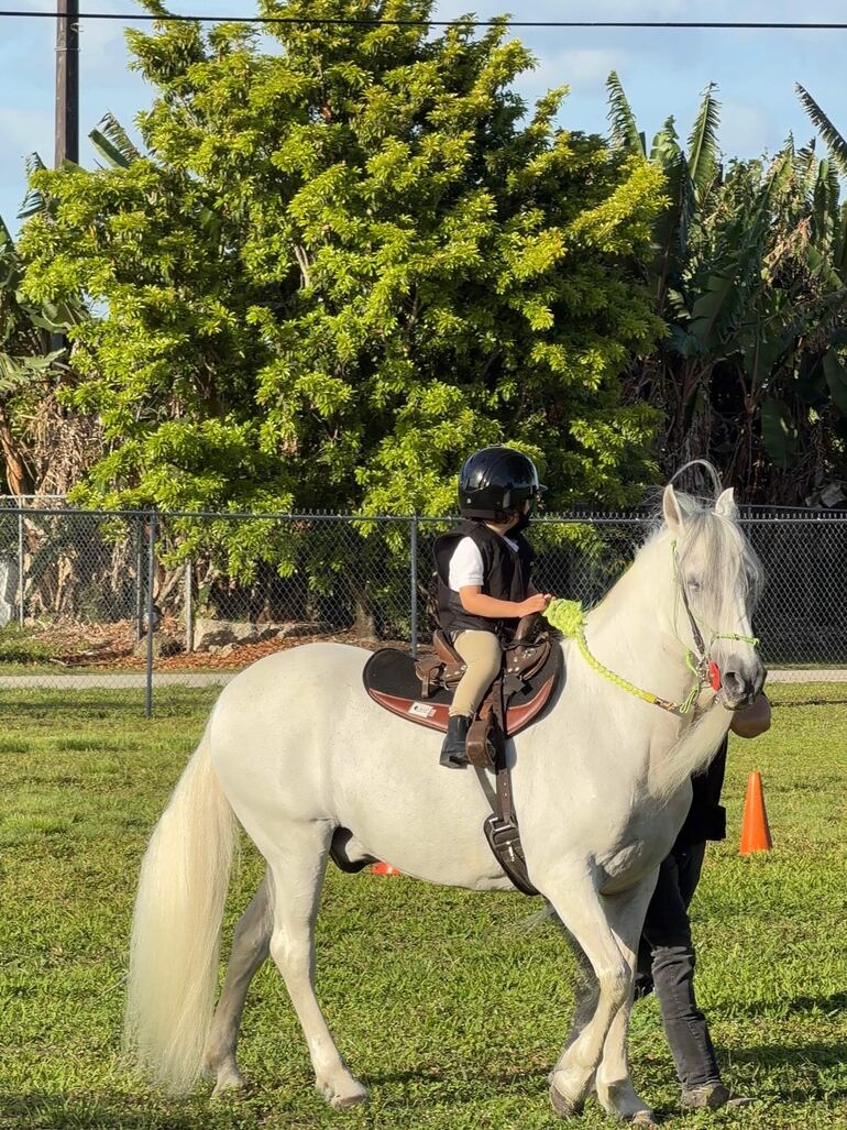 Niño con casco negro y camisa blanca montando un caballo blanco, mientras un adulto lo sostiene a su lado en un entorno verde.