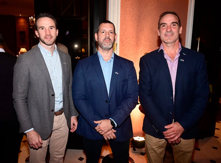 Tres hombres en blazers interactuando en un evento social, uno con expresión seria, otro sonriente y el tercero con postura relajada.