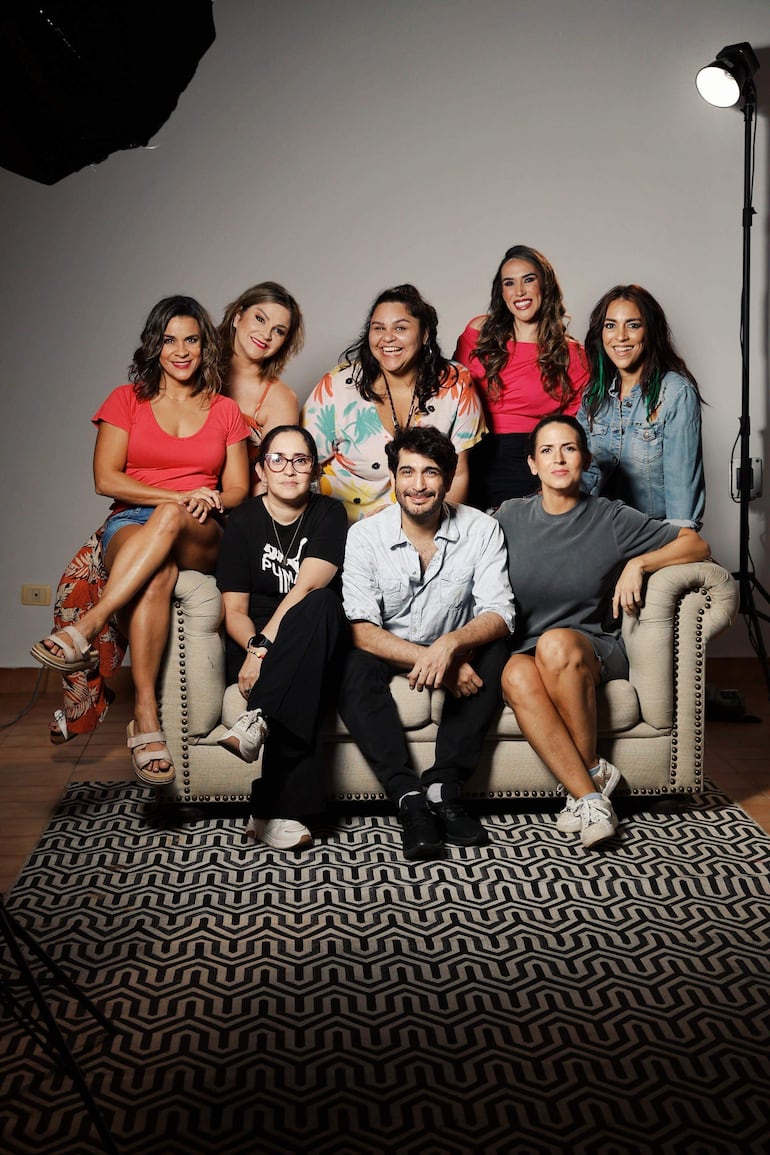Diego Mongelós rodeado de siete actrices felices en un sofá, iluminados suavemente en un fondo neutro.