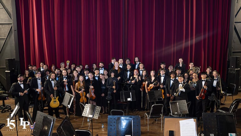 Cincuenta músicos vestidos de negro en el escenario con instrumentos, listos para una actuación en un ambiente formal con cortina roja.