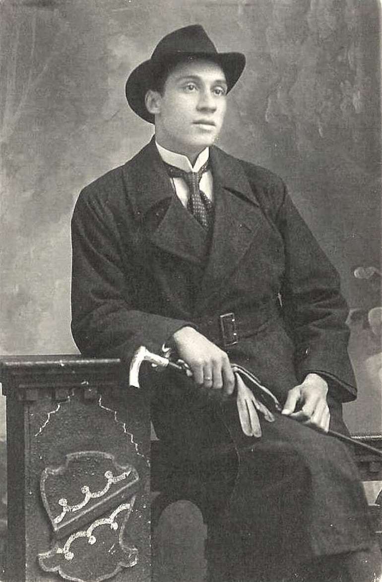 Joven hombre con abrigo oscuro y sombrero de ala ancha, sentado con expresión seria en un estudio difuso.