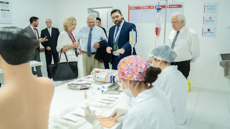 Mujer en blusa blanca y bufanda roja observa a dos profesionales en batas blancas, rodeados de instrumentos médicos en ambiente bien iluminado.
