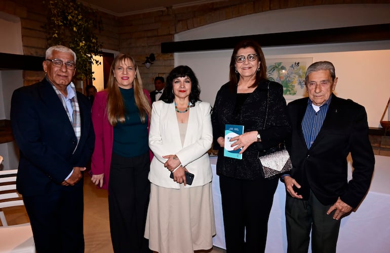 Zully Vera de Molinas con abrigo fucsia en evento, rodeada de cuatro personas posando formalmente en un ambiente decorado con piedra.