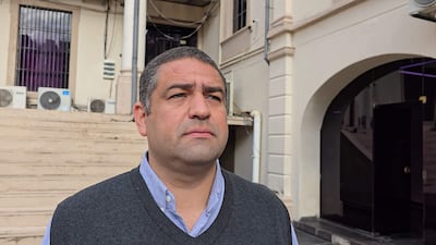 José Ramón Rodríguez Maciel con chaleco gris y camisa celeste, luciendo serio frente a estructuras arquitectónicas y escaleras.